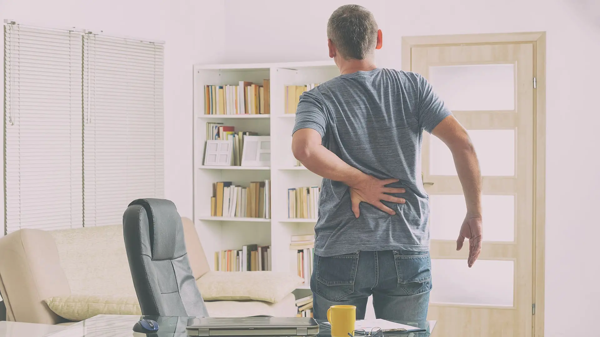 Chronic Back pain Treatment. A man standing next to a desk with his hand on his lower back indicating pain
