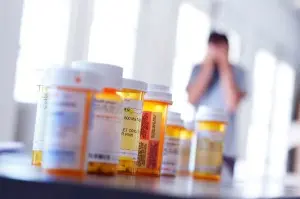 Closeup of a row of prescription pain pill bottles, while a man holds his face in his hands in the background